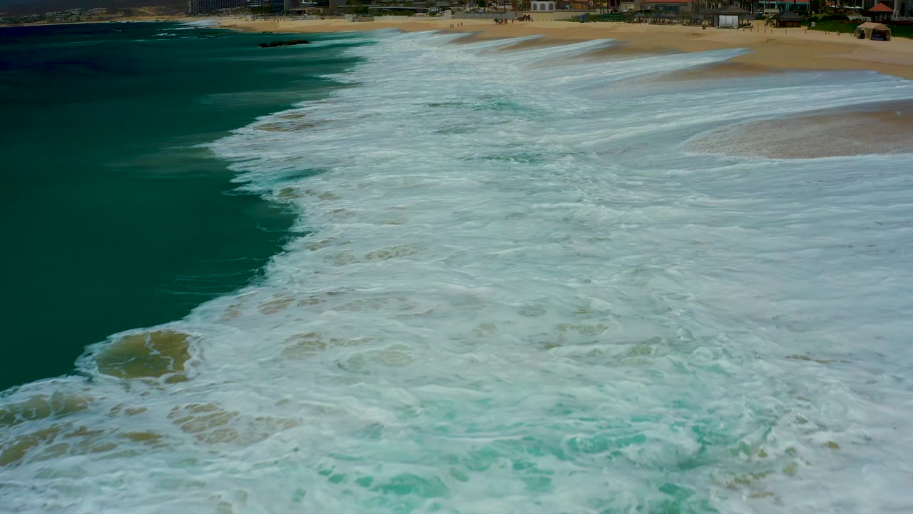 Aerial view over Dreams Los Cabos surf where white foam fills most of the frame and the sandy beach appears across the top right, with no visible people.