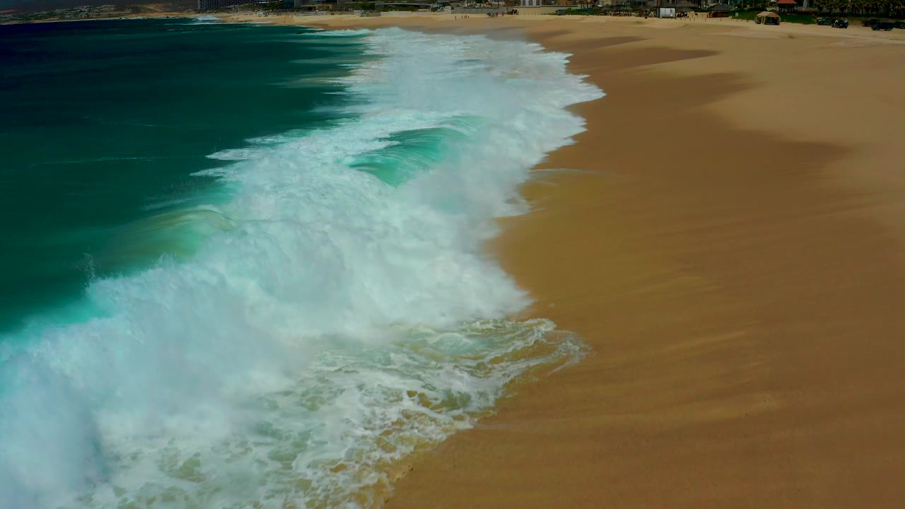 Aerial frame from Dreams Los Cabos with a larger band of white foam moving diagonally over wet golden sand, turquoise water at left, and no visible people.