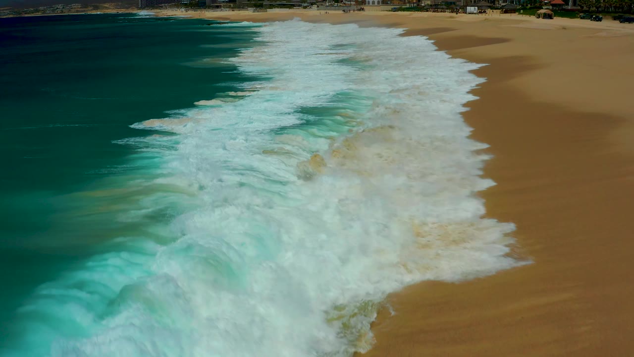 Dreams Los Cabos aerial view with foamy surf spreading over the beach, darker turquoise water along the left edge, exposed golden sand at right, and no visible people.