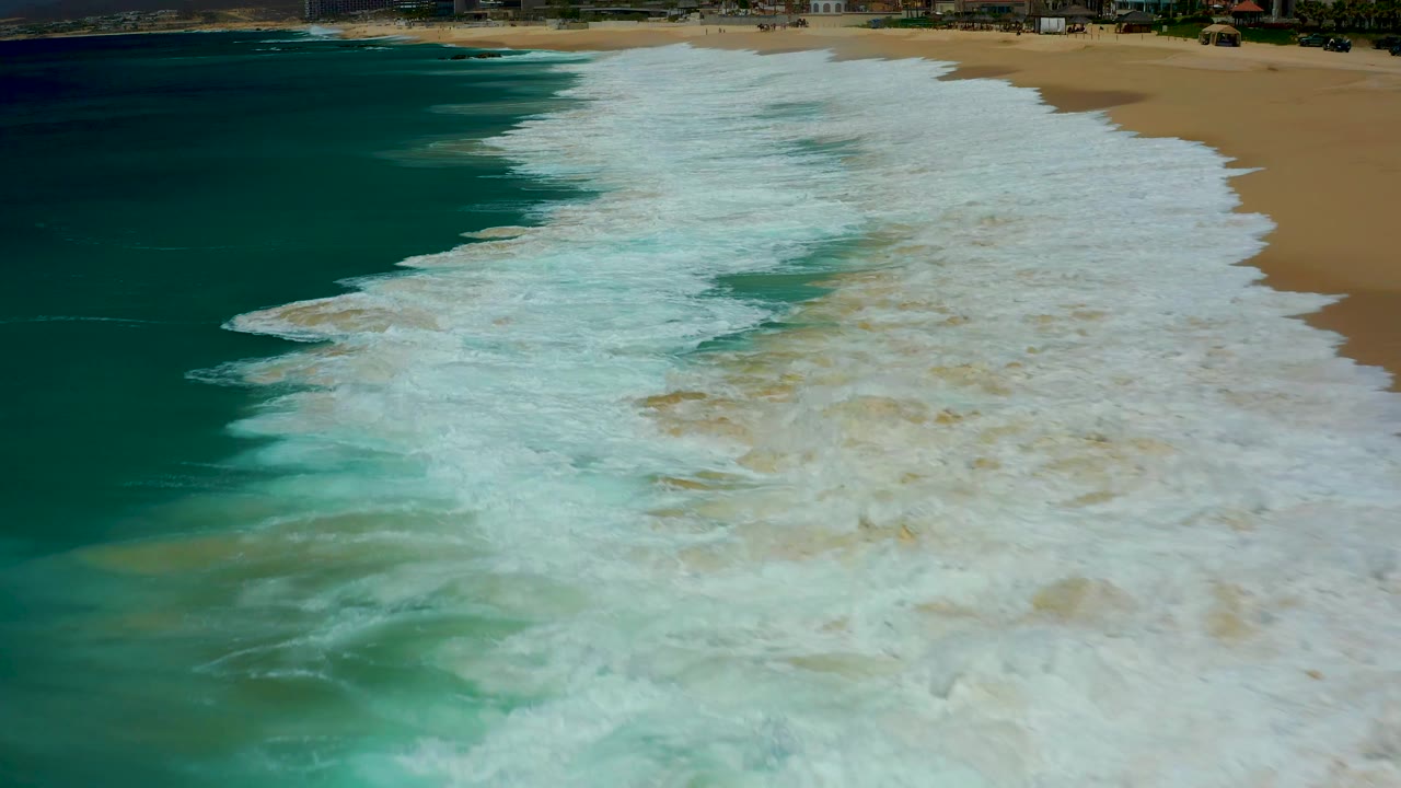 Aerial shot at Dreams Los Cabos where a broad white surf band fills the center, dark teal ocean remains on the left, golden sand borders the right side, and no visible people appear.