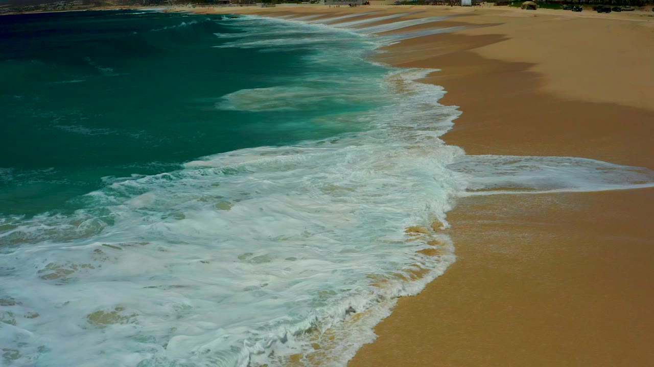 Elevated view of turquoise surf washing onto the sandy shore at Dreams Los Cabos, Mexico, with no visible people.