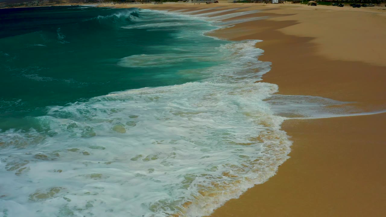 Foamy surf reaches across the beach while open dry sand remains on the right at Dreams Los Cabos, Mexico, with no visible people.