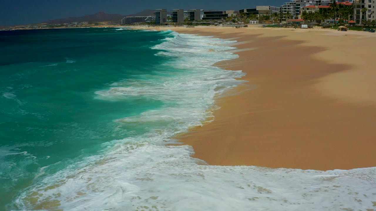 At Dreams Los Cabos in Baja California Sur, turquoise surf approaches a broad sandy shoreline with beachfront buildings along the top of the frame and no visible people.