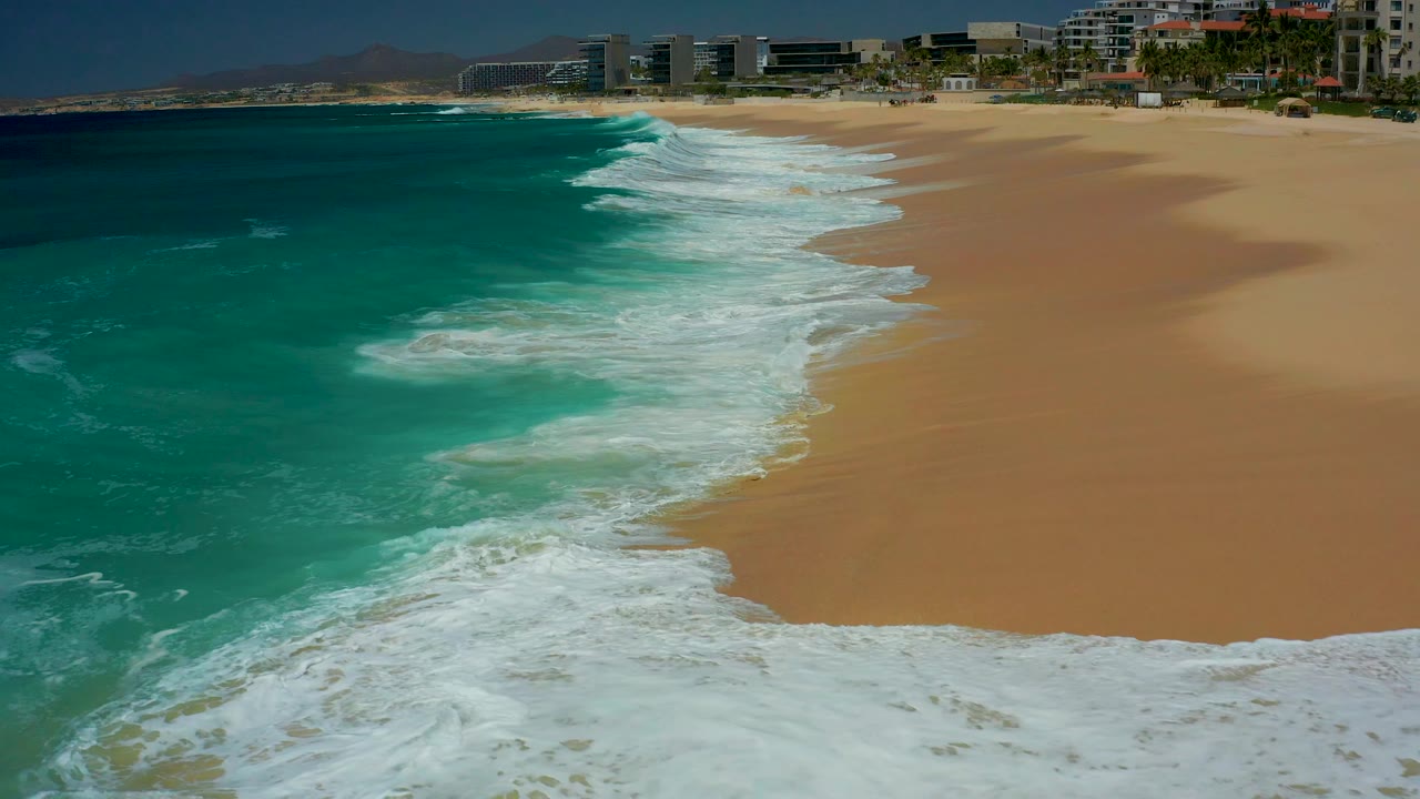 White foam expands along the Dreams Los Cabos shoreline while clear turquoise water holds the left side of the frame, with distant buildings and no visible people.