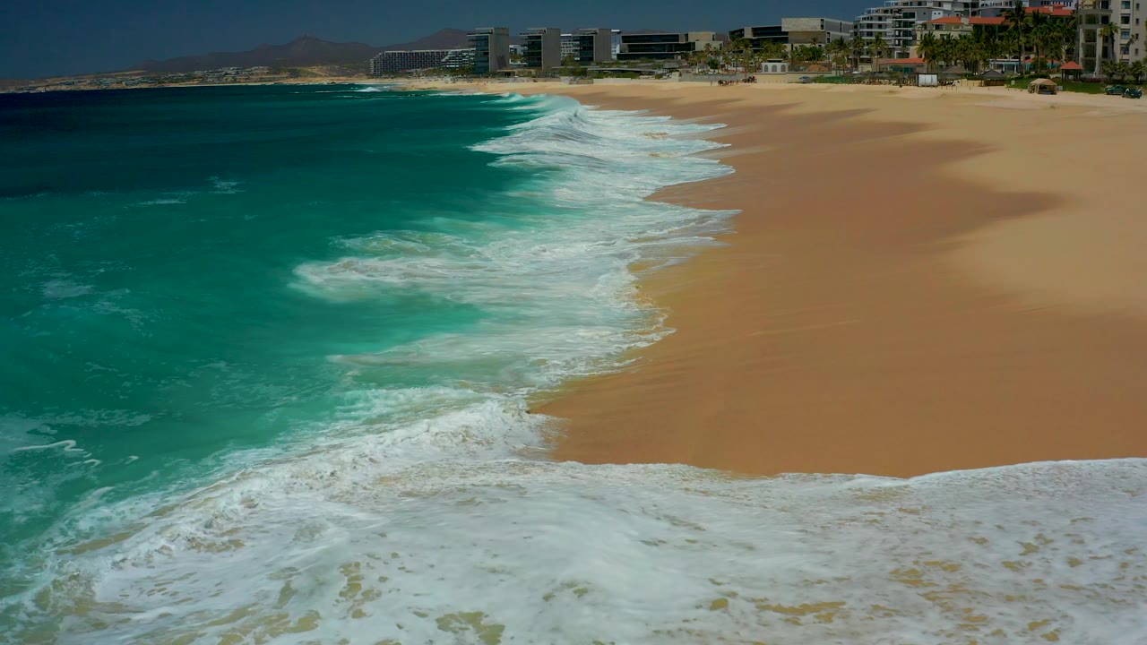 A thicker sheet of white surf reaches the lower frame edge at Dreams Los Cabos, with open sand to the right, turquoise sea to the left, and no visible people.