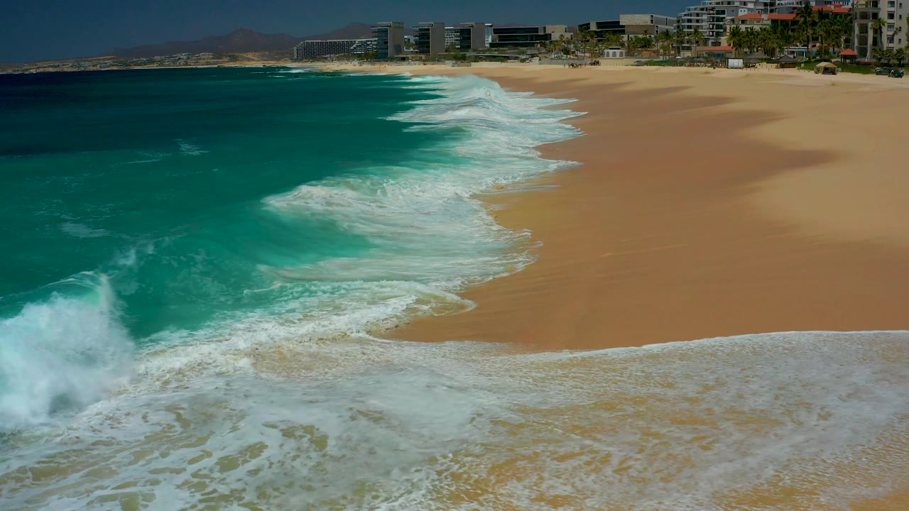 Foamy water fills the lower foreground at Dreams Los Cabos, the shore curves toward distant beachfront buildings, and the beach remains empty with no visible people.
