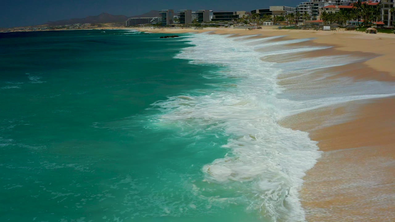 At Dreams Los Cabos in Baja California Sur, turquoise water and white foam angle onto tan sand with distant beachfront buildings along the upper edge and no visible people.