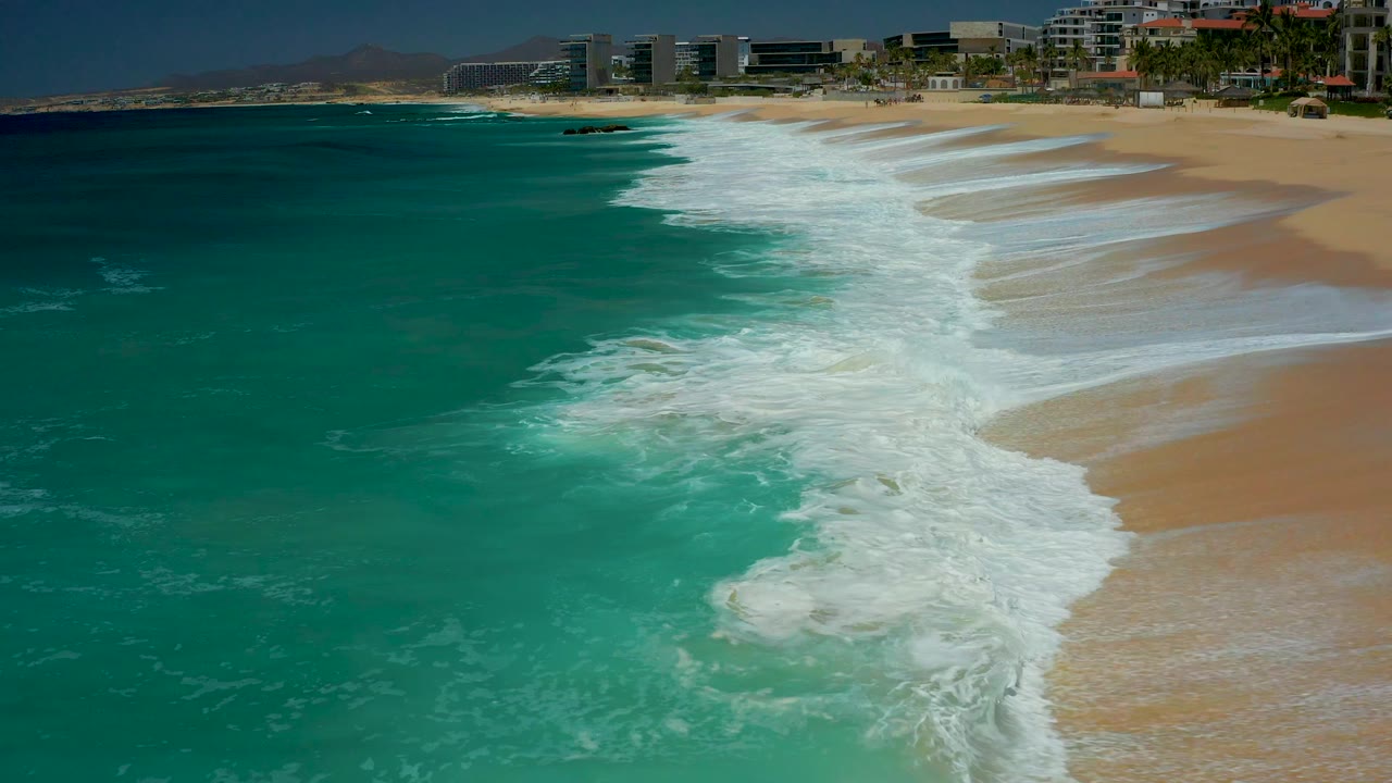 At Dreams Los Cabos in Baja California Sur, a wider band of white foam spreads across the center shoreline while turquoise water stays to the left and no visible people appear on the beach.
