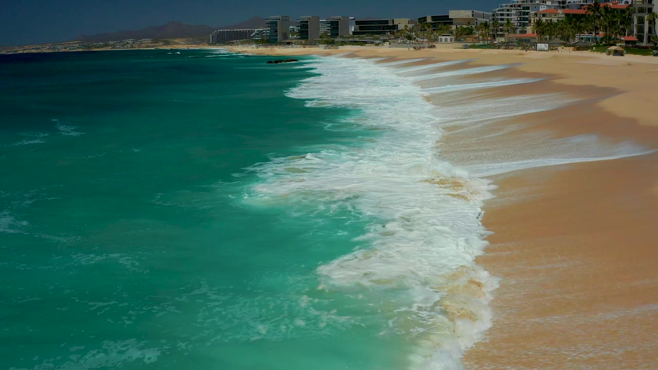 At Dreams Los Cabos in Baja California Sur, the surf line curves down the center of frame, leaving wet sand patterns to the right and open turquoise water to the left with no visible people.