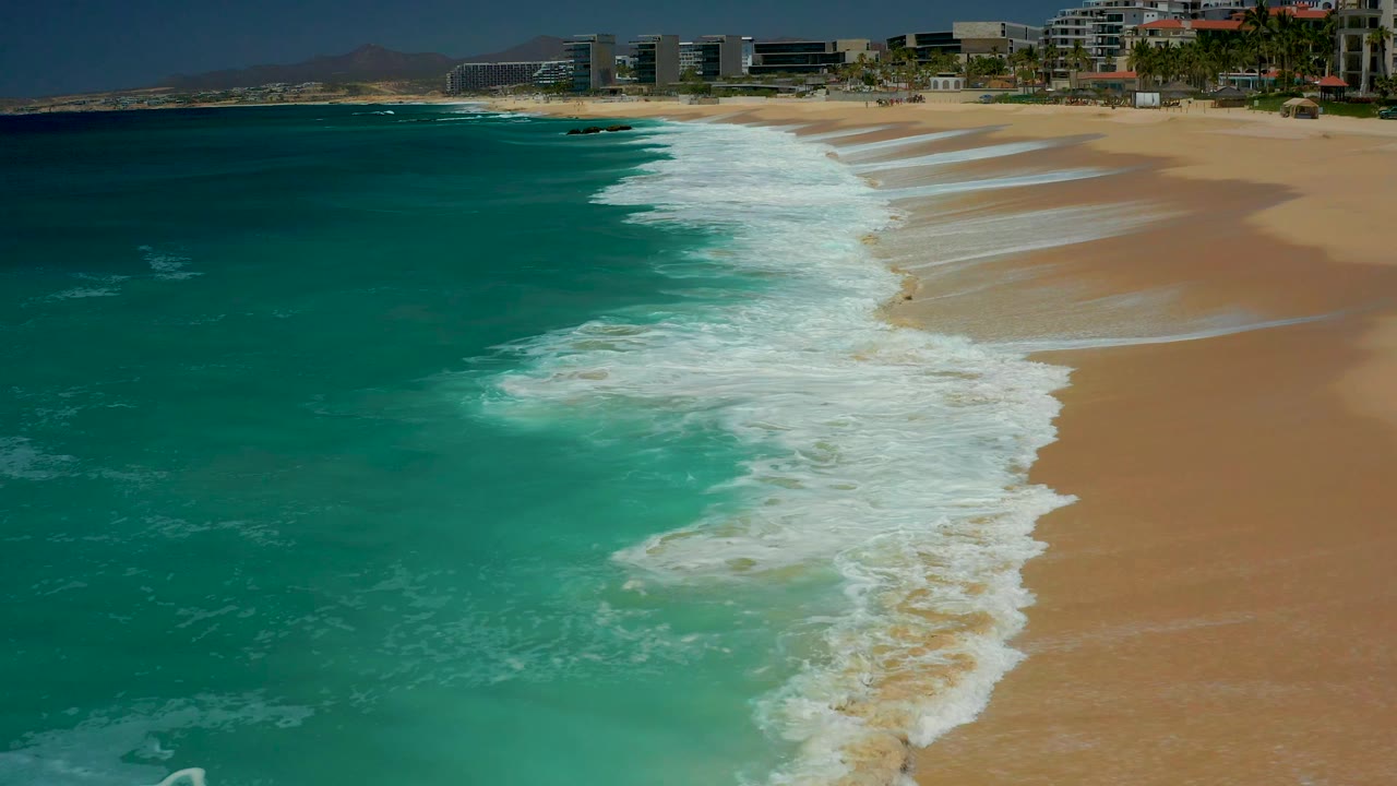 At Dreams Los Cabos in Baja California Sur, white wave wash stretches from the lower center toward the distant beach, bordered by teal ocean and empty tan sand with no visible people.
