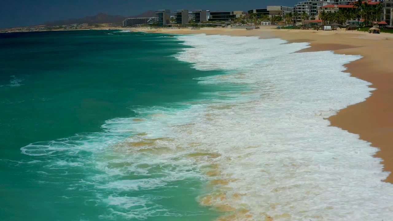 Aerial view over Dreams Los Cabos beach as a broad sheet of white foam pushes across tan sand, with turquoise water to the left and no visible people on the shore.