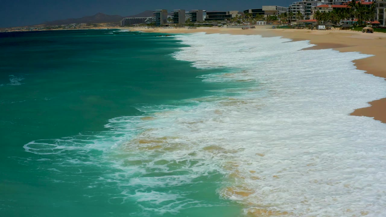 Aerial shoreline frame at Dreams Los Cabos with breaking white waves filling the right side, deeper turquoise water on the left, distant buildings along the beach, and no visible people.