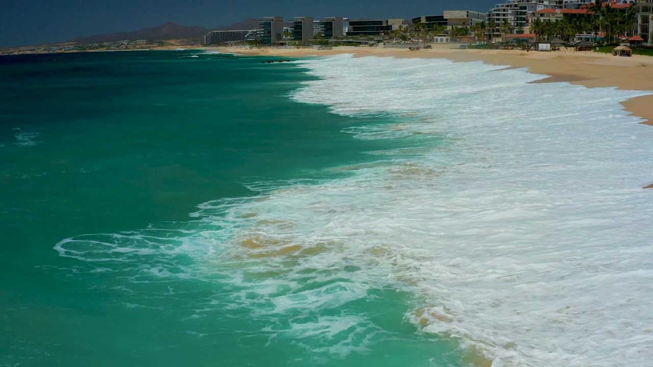 Aerial frame from Dreams Los Cabos showing a rounded patch of foam in green-blue water, the sandy beach along the right edge, distant resort structures, and no visible people.