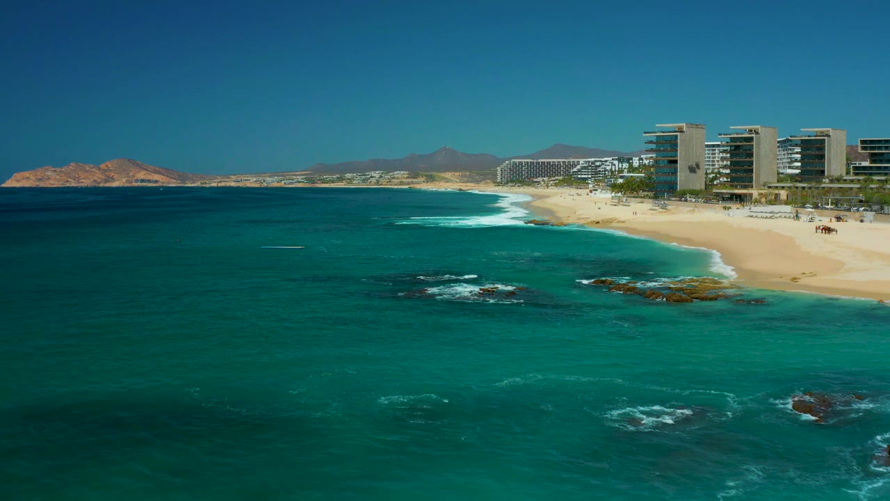 One second into the clip, Dreams Los Cabos beachfront buildings sit along the right edge while white waves curve across the sand and rocks show through the water, with no visible people.