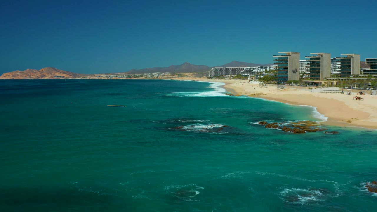 At 0:02, the ocean foreground dominates the frame, with a thin band of white surf reaching the sand below the Dreams Los Cabos buildings and no visible people.