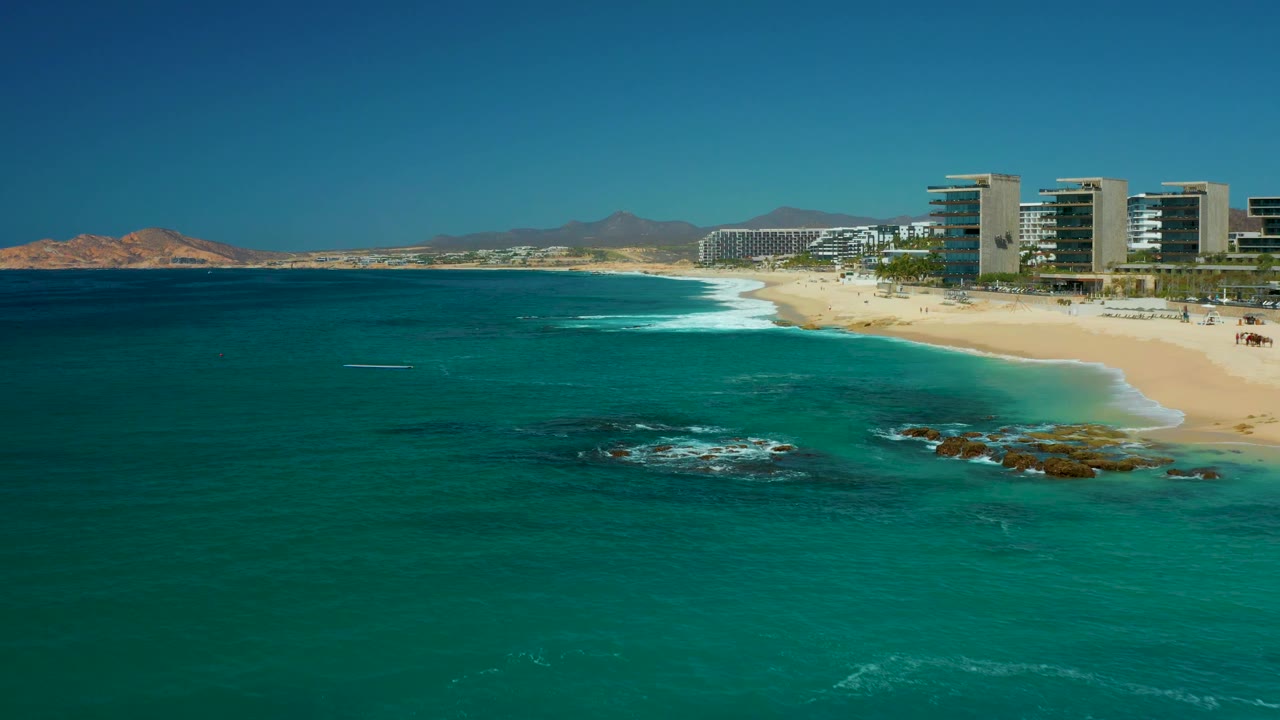Beachfront buildings above a sandy shore with turquoise water and rocky surf.