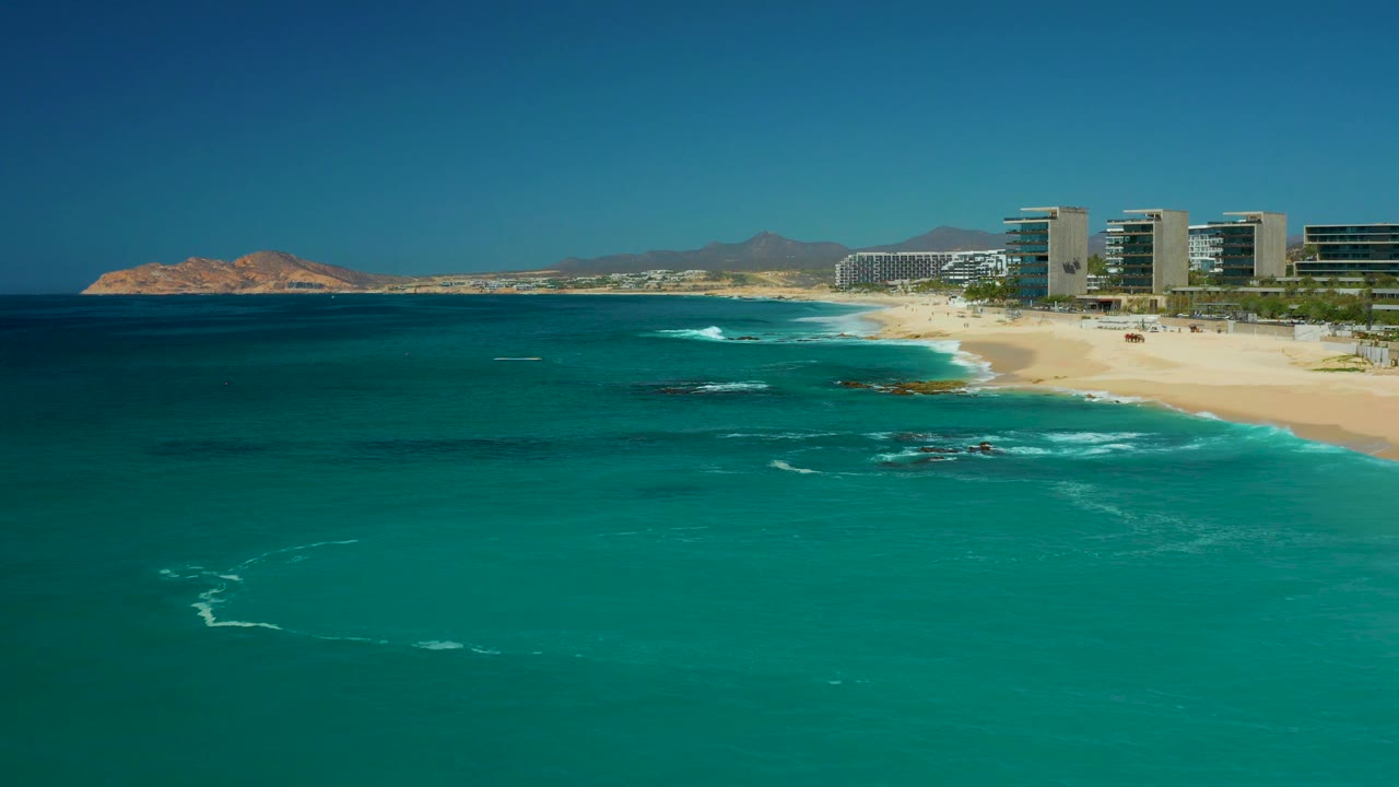 At Dreams Los Cabos, Mexico, the frame shows open turquoise water, a thin line of white surf near shore, resort buildings along the right side, distant rocky landforms, and no visible people.