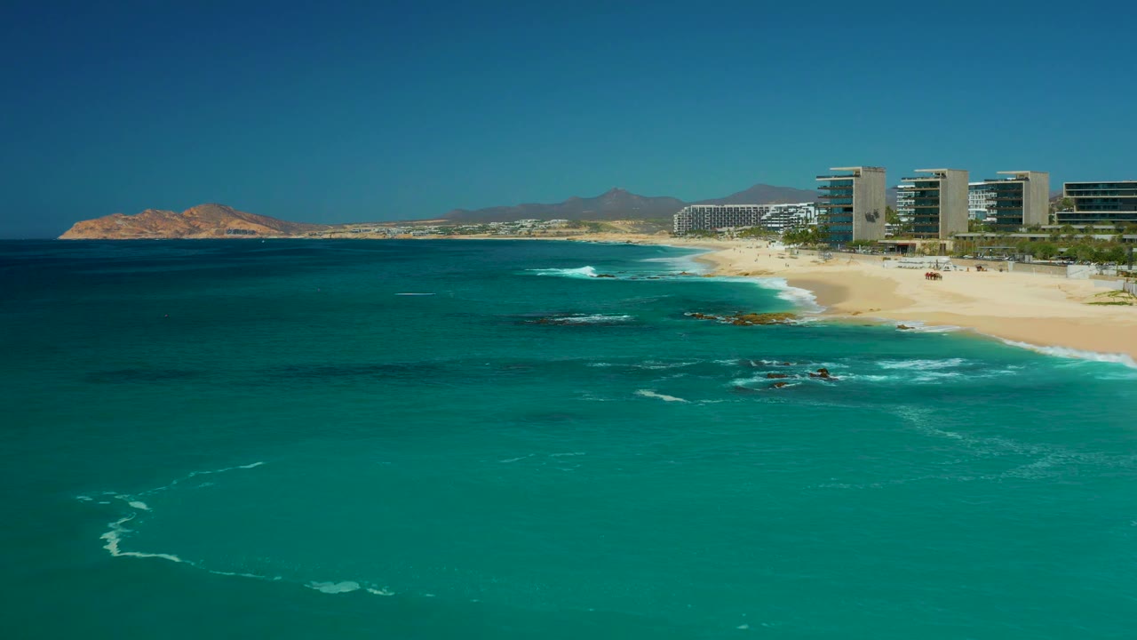 The Dreams Los Cabos shoreline remains in a wide view, with darker reef or rock patches under the water, white wave foam near the sand, buildings on the right, and no visible people.