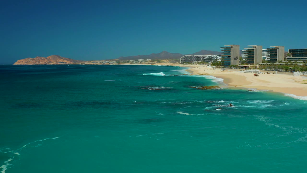 A wider section of the Dreams Los Cabos coast shows turquoise water across the foreground, breaking waves over dark rocks, sandy beach to the right, resort buildings behind it, and no visible people.