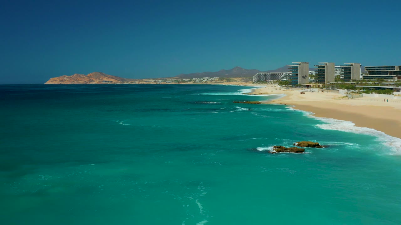 Opening frame over Dreams Los Cabos, Baja California Sur, with turquoise water in front, small waves near the sand, offshore rocks, beachfront buildings in the distance, and no visible people.