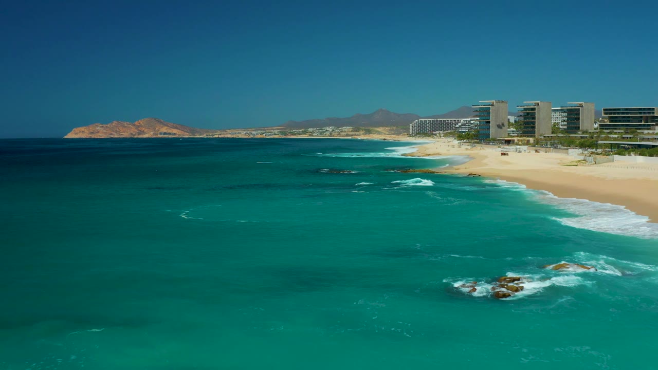Aerial beach view with dark turquoise water, breaking foam around rocks, and buildings along the sand.