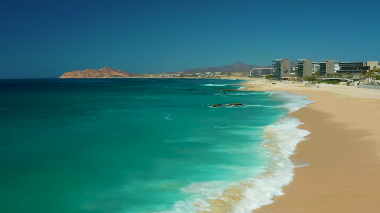 Aerial view at Dreams Los Cabos in Baja California Sur showing turquoise water, white surf on pale sand, beachfront buildings, distant hills, and no visible people.