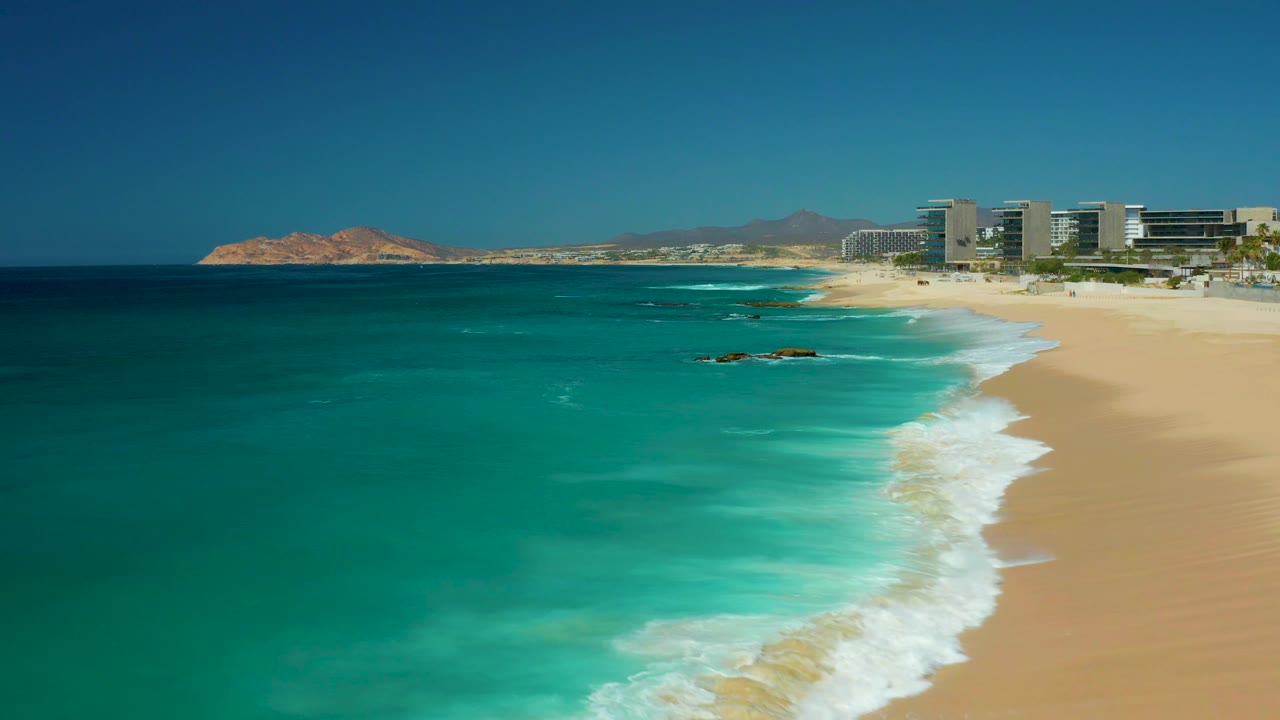 White waves curl along a sandy shoreline with turquoise water and resort buildings in the distance.
