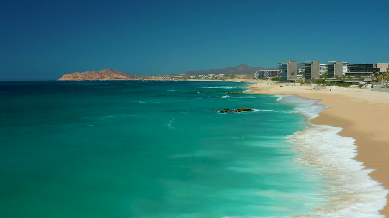 At 0:02, the aerial composition shifts toward open water, with white surf streaking along the right edge of the Dreams Los Cabos beach, distant hills, buildings, and no visible people.