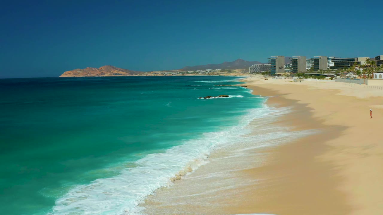 At 0:01, the surf line has advanced across the sand while the Dreams Los Cabos shoreline and distant buildings remain clear, with no visible people.