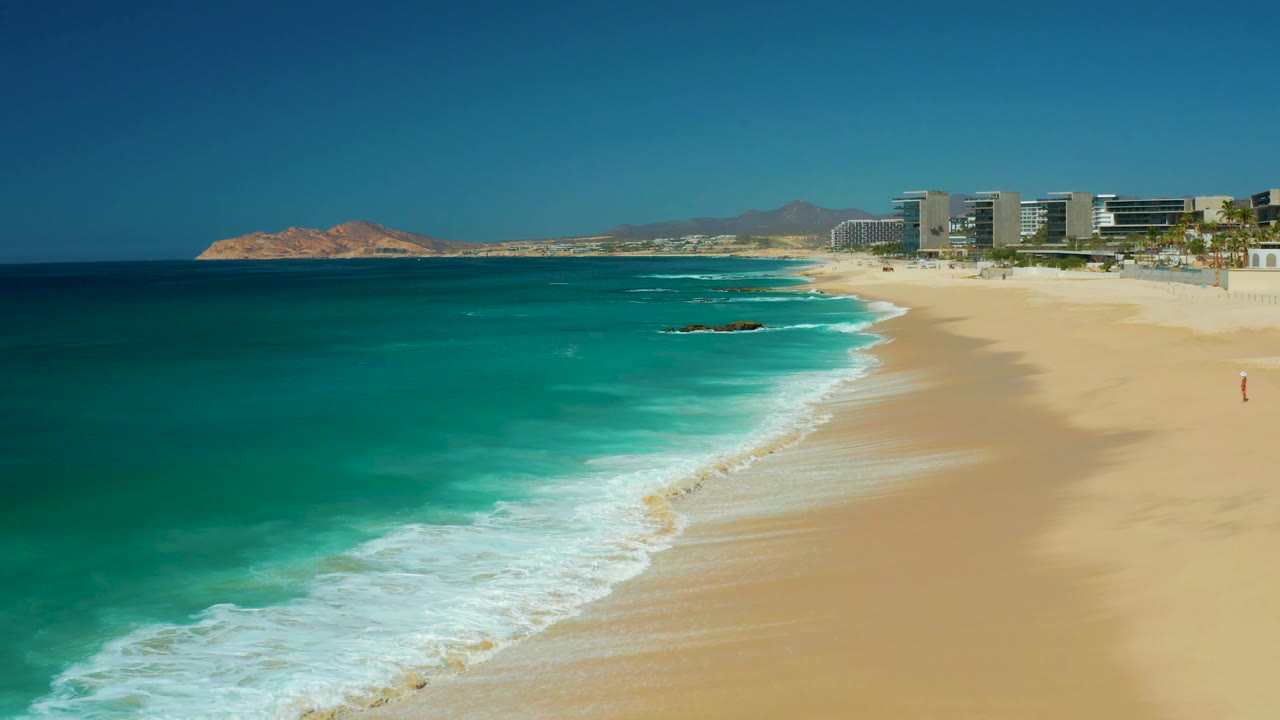 At 0:03, turquoise water and white foam fill the left and lower frame while the broad Dreams Los Cabos beach remains empty, with no visible people.