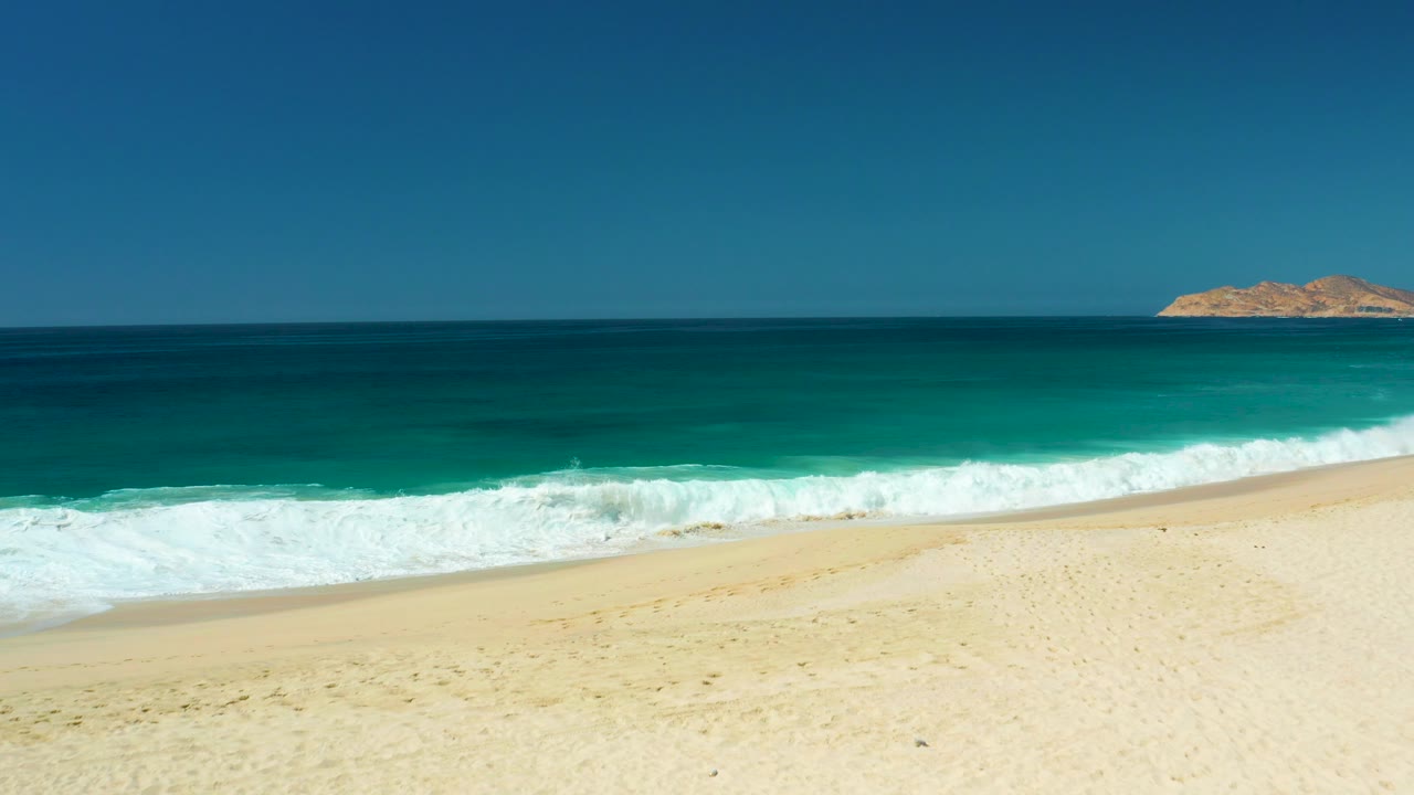 At Dreams Los Cabos in Baja California Sur, white surf rolls across pale sand below turquoise and deep blue water, with a small landmass on the horizon and no visible people.