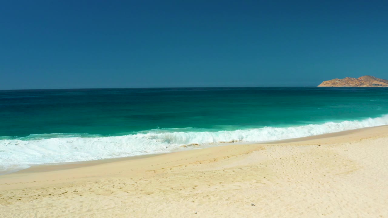 Beach surf advancing along pale sand with turquoise water and a distant rocky landmass.