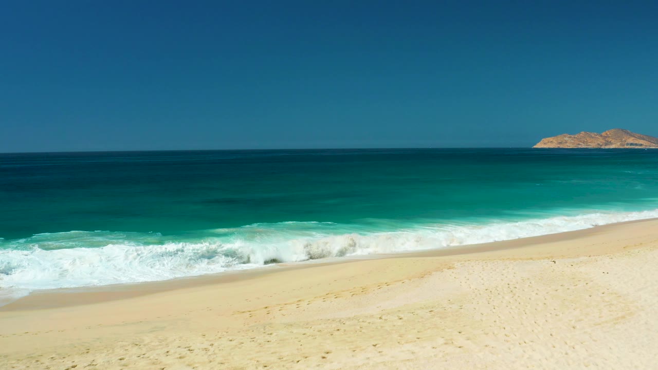 At Dreams Los Cabos, a wider band of white foam breaks toward the sandy shore, with the ocean layered from turquoise near shore to dark blue offshore and no visible people.