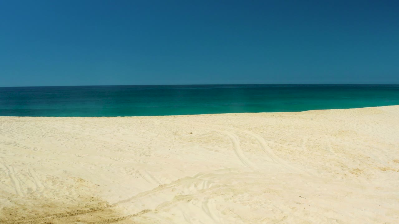 Wide view at Dreams Los Cabos in Baja California Sur with pale sand in the foreground, a calm blue ocean band, clear sky, and no visible people.