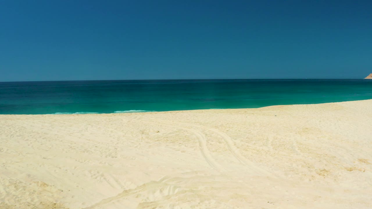 The 0:02 frame holds a wide sandy shoreline at Dreams Los Cabos, with deep blue water beyond the pale beach and no visible people.