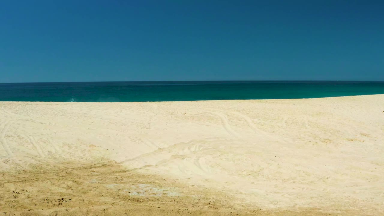 Opening frame of Dreams Los Cabos beach with pale sand in front, a dark blue ocean band at the horizon, clear sky above, and no visible people.