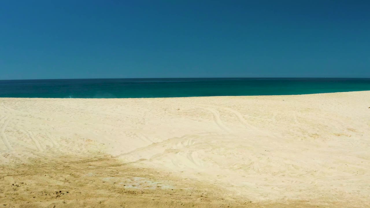 Wide frame at Dreams Los Cabos showing bright pale sand, turquoise water near shore, deeper blue water beyond it, clear sky, and no visible people.