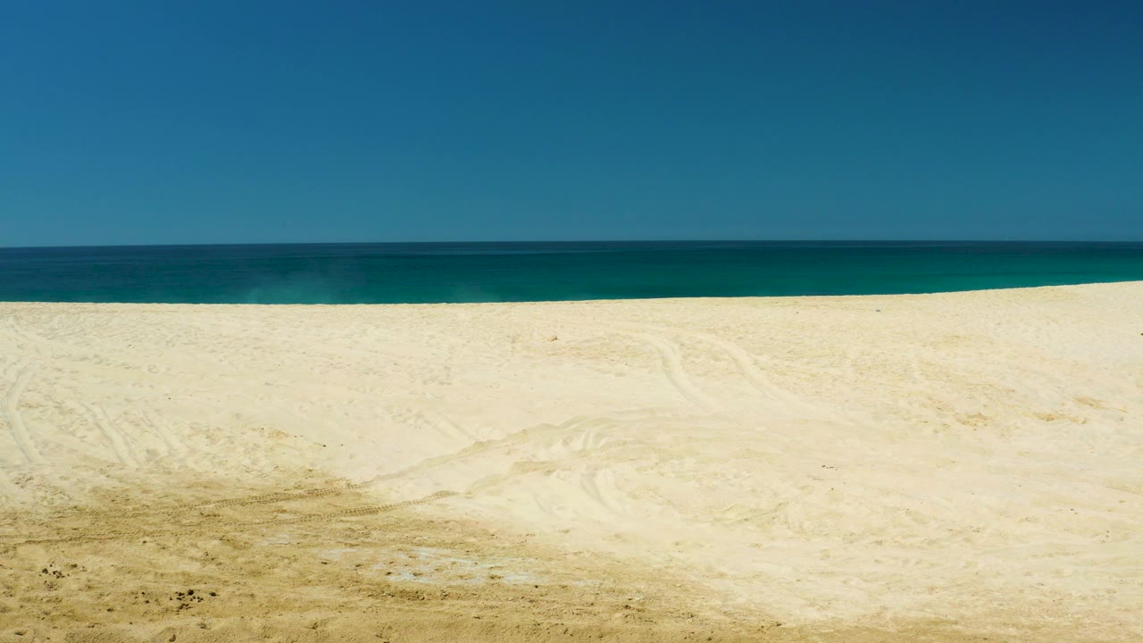 Sunlit Dreams Los Cabos shoreline frame with textured sand across the foreground, a narrow ocean band in the distance, open blue sky, and no visible people.