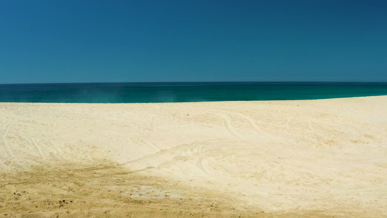 Middle frame of the Dreams Los Cabos beach view with a low straight horizon, broad pale sand, blue-green water, clear sky, and no visible people.