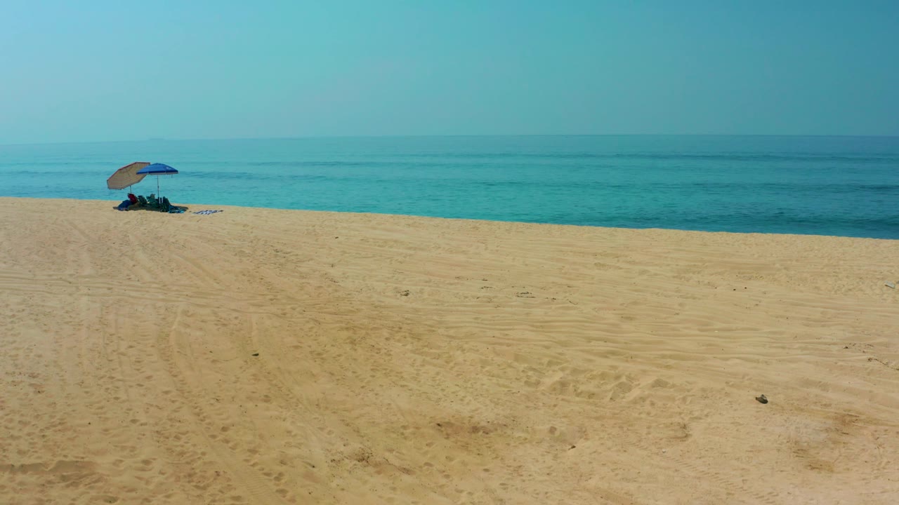 Shipwrecks Los Cabos beach view with broad sand in front, a blue umbrella and small people near the waterline, and calm blue ocean under a pale sky.
