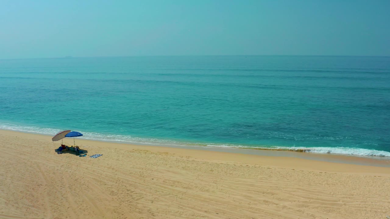 At Shipwrecks Los Cabos, a high wide view shows open sand, a blue umbrella near the shoreline, turquoise ocean beyond, and no visible people.