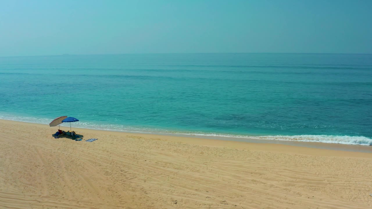 At Shipwrecks Los Cabos, the umbrella appears farther left in the wide sandy frame, with calm turquoise water and a pale sky above, and no visible people.