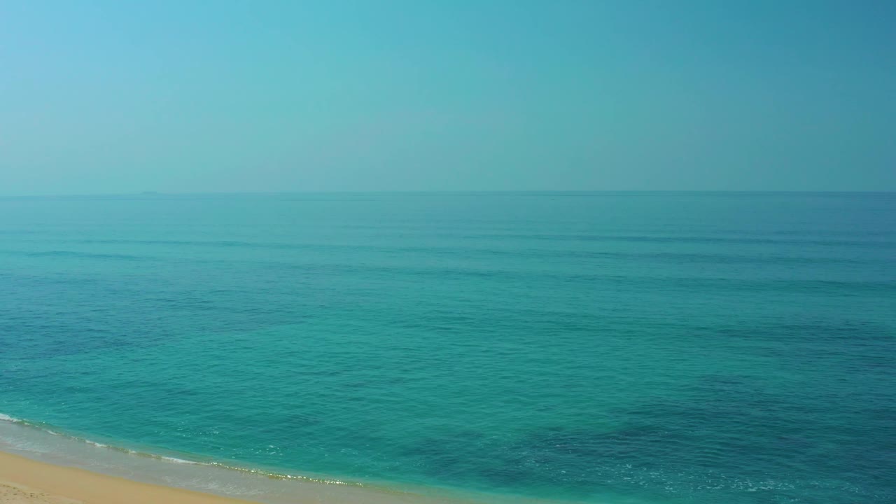 Aerial frame over Shipwrecks Los Cabos showing calm turquoise water, faint wave texture, a slim beach strip, and no visible people.