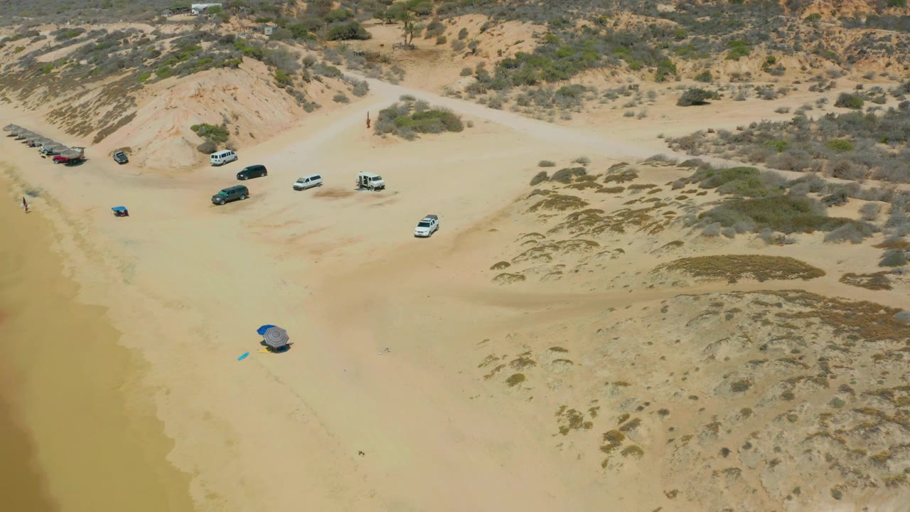 The next Shipwrecks Los Cabos frame keeps the parked vehicles near the upper sand while the blue shelter sits closer to the shoreline edge, with dune vegetation across the right side and no visible people.