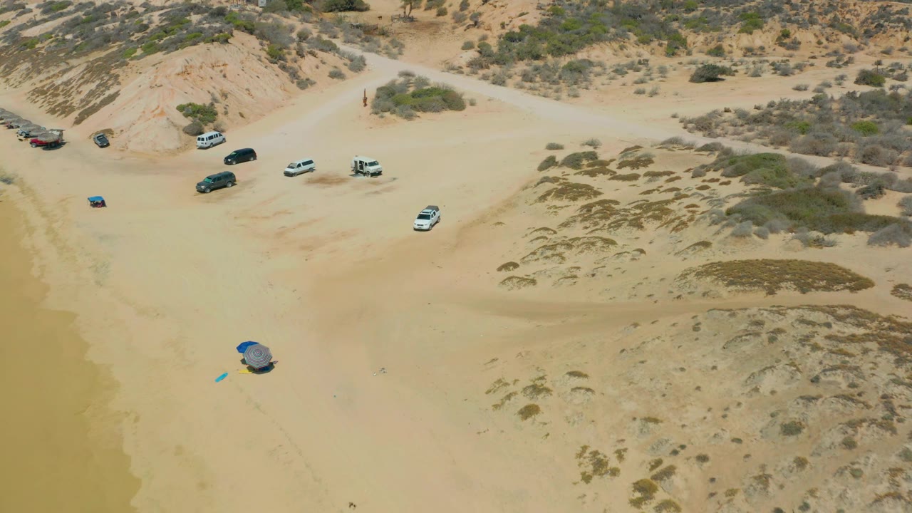 A high drone view at Shipwrecks Los Cabos shows several vehicles spread along the upper beach, a blue shelter on the open lower sand, patchy dune vegetation, and no visible people.