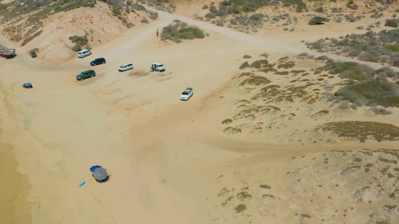 At mid-clip, the Shipwrecks Los Cabos beach view places the blue shelter near the lower left, several vehicles along the upper beach, scrubby dunes to the right, and no visible people.