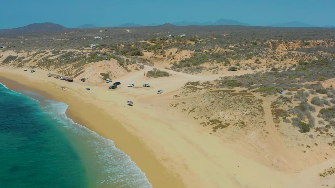 Aerial view of turquoise water, sandy beach, dunes, and parked vehicles.
