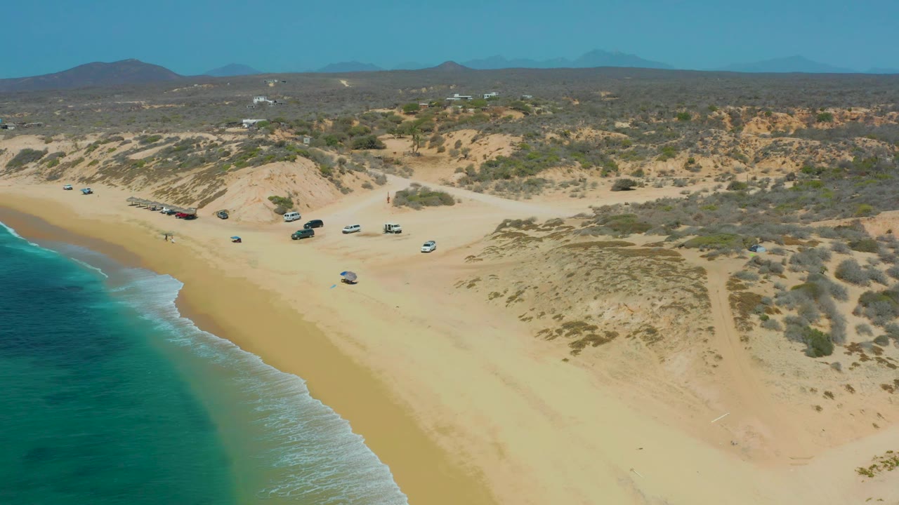 Aerial frame at 0:01 over Shipwrecks Los Cabos showing the surf line edging the beach, several parked vehicles beside the dunes, low vegetation, distant hills, and no visible people.