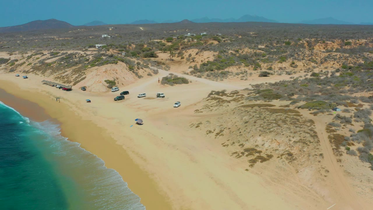 Mid-clip aerial frame over Shipwrecks Los Cabos with ocean foam at left, a broad sandy beach, multiple vehicles parked near a dune access path, scrub vegetation, hills on the horizon, and no visible people.