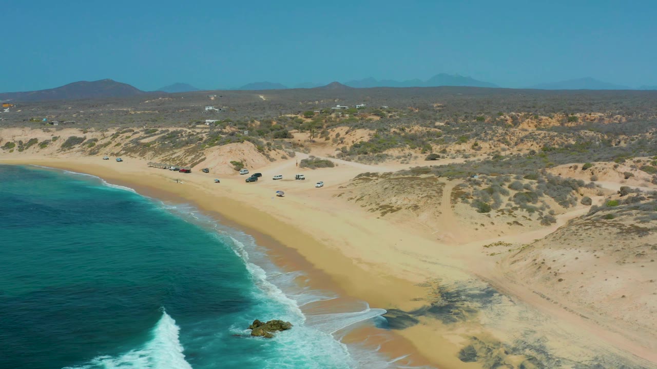 Aerial frame at Shipwrecks Los Cabos with the surf line slightly farther along the beach, a small rock in the breaking water, dry dunes beyond the sand, parked vehicles near the shore, and no visible people.